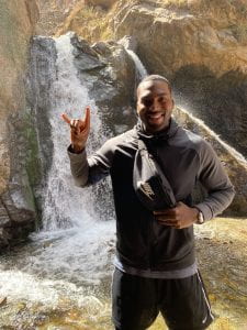 Black man smiles at camera in front of waterfall