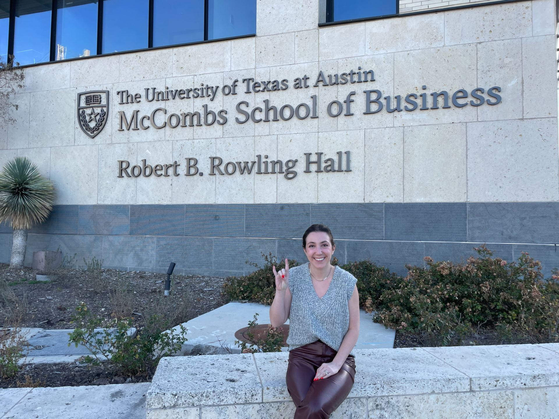 Marion sits outside of a Texas McCombs sign in thr front of our building. The sign reads "The University of Texas at Austin: McCombs School of Business - Robert B. Rowling Hall"